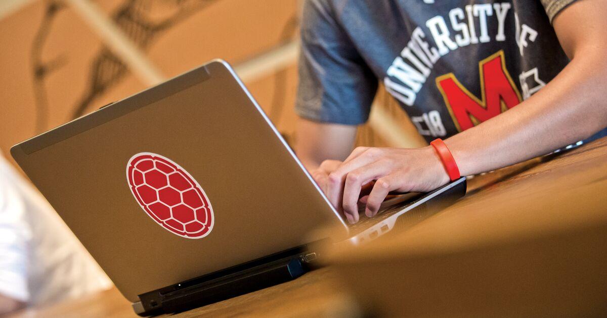 male student using his laptop on a table in Starbucks with another student in the background, shell sticker on the laptop.