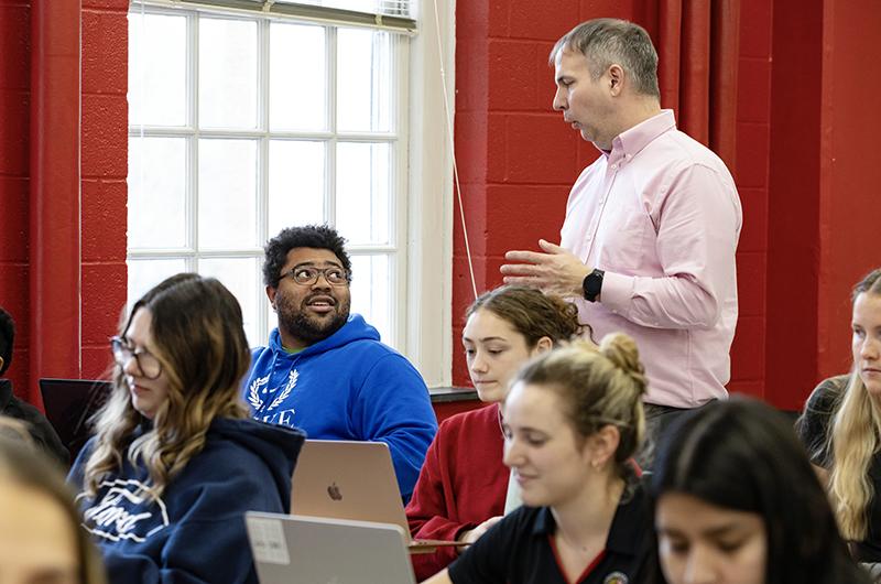 A professor standing speaks to students sitting in a classroom with bright red walls.