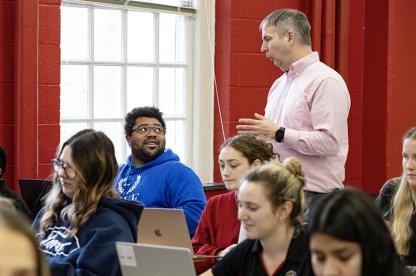 A professor standing speaks to students sitting in a classroom with bright red walls.
